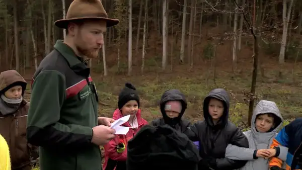 Ein Förster mit braunem Hut erklärt Kindern im Wald etwas. Sie sind von Hütten aus hierher gekommen, um mehr über die Natur zu lernen.