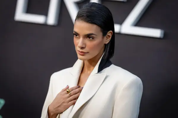 Laysla de Oliveira, elegant in a white blazer, poses with a hand on her chest, adorned with a striking gold ring.