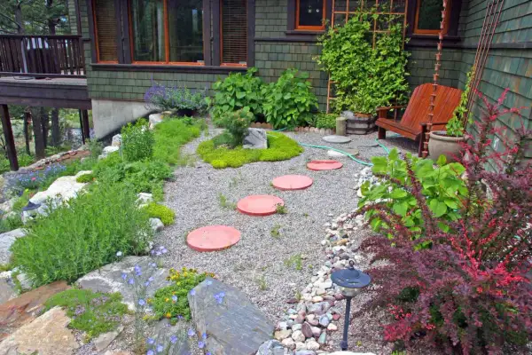 Inexpensive stepping stone walkway ideas: red circular pavers create a path through a gravel garden with lush greenery and a wooden chair.