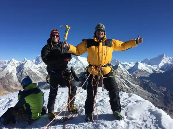 Dwóch alpinistów na szczycie Mera Peak, jeden z czekanem, drugi z kciukiem w górę. W tle ośnieżone góry.