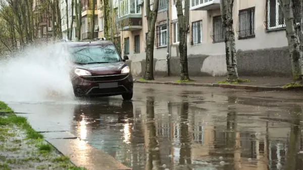 Czerwony samochód wpada w kałużę, tworząc fontannę wody. To przykład aquaplaningu, czyli utraty przyczepności na mokrej nawierzchni.