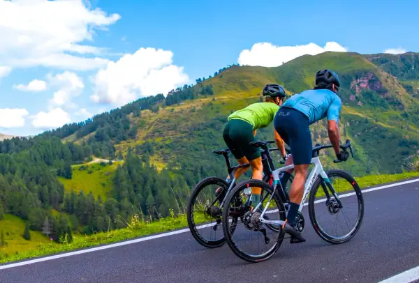 Dwóch kolarzy w sportowych strojach pokonuje górską trasę. Ich pozycja na rowerze szosowym jest aerodynamiczna, gotowi na wyzwanie.