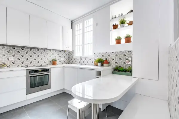 Cuisine moderne aux meubles blancs épurés, rehaussée par une crédence aux motifs géométriques gris et blancs. Une table blanche intégrée et des plantes apportent une touche de fraîcheur.