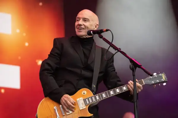 Łysy mężczyzna w czarnym garniturze gra na gitarze elektrycznej, uśmiechając się. To jest jego chwila, bez żalu.