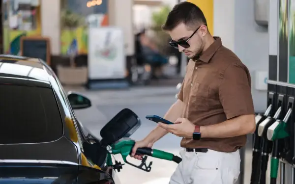 Un homme fait le plein de sa voiture à une **téléphone station essence**, consultant son smartphone.