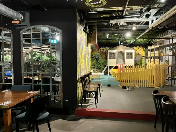 Warszawska restauracja z salą zabaw dla dzieci. Widać stoliki, krzesła i kolorowy plac zabaw z domkiem i zjeżdżalnią.