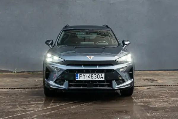 Szary Formentor Cupra z włączonymi światłami, zaparkowany przed betonową ścianą.