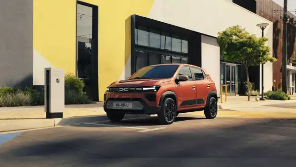 En orange Dacia står parkerad vid en laddstation framför en modern byggnad. Är Dacia bra eller dålig? Denna ser lovande ut!