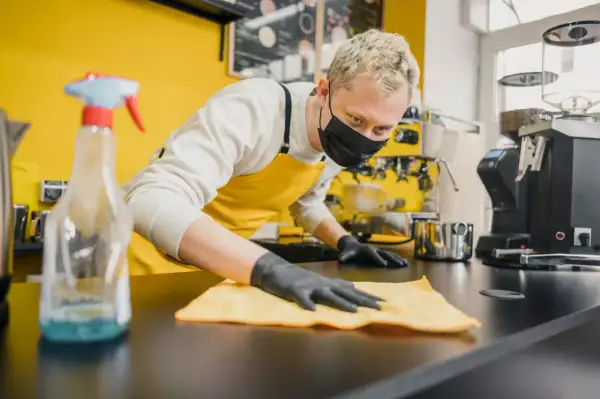 Barista con mascherina e guanti effettua la pulizia della cucina del ristorante, igienizzando il bancone con un panno giallo.