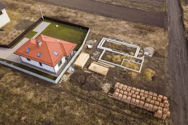Zabudowa zagrodowa w budowie: gotowy dom z czerwonym dachem, fundamenty nowego budynku, drewno i palety cegieł.