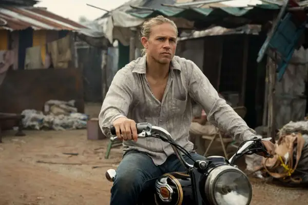 Charlie Hunnam kör motorcykel genom ett slumområde, med en blick av beslutsamhet.