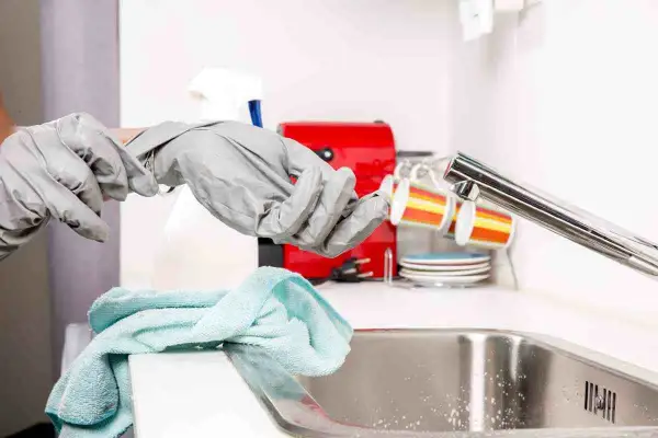 Mani con guanti grigi si preparano a sturare il lavandino, con un panno azzurro e una macchinetta del caffè rossa sullo sfondo.