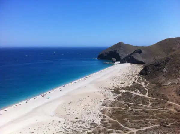 Playa de arena blanca y mar azul intenso, con acantilados rocosos y vegetación árida. Un velero navega en la distancia, un capricho del paisaje.