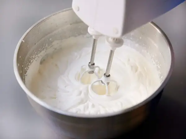 Schlagobers wird mit einem Handmixer in einer Edelstahlschüssel aufgeschlagen. Die cremige Masse ist bereit für süße Köstlichkeiten.