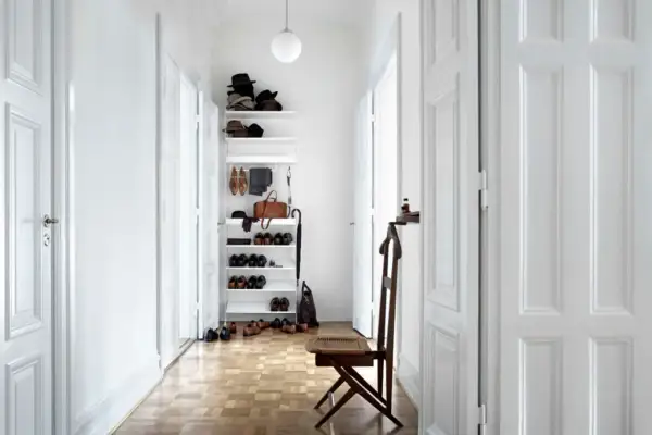 Helle Türen und ein schmaler Flur optisch breiter wirken lassen. Ein weißes Regal mit Hüten, Schuhen und einer Tasche steht im Flur.