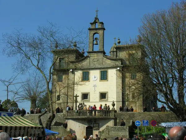 Edificio religioso con campanario y cruz. Gente en el balcón y en la plaza. Un árbol desnudo en primer plano.
