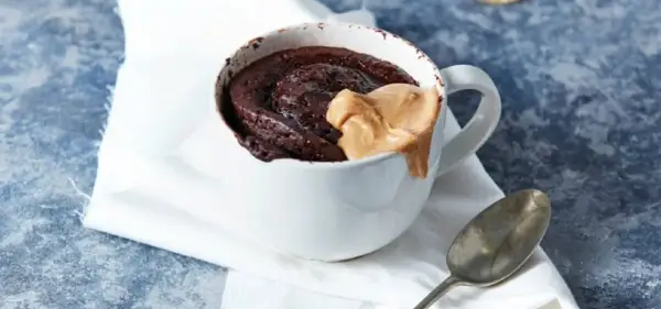 Schokoladiger high protein tassenkuchen mit Erdnussbutter-Topping in einer weißen Tasse, daneben ein Löffel.