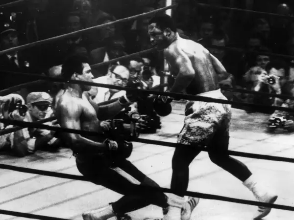 Joe Frazier fights in the ring, delivering a powerful blow as his opponent stumbles. The crowd watches intently.