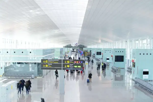 Jasny, nowoczesny terminal lotniska w Hiszpanii. Pasażerowie z bagażami przemieszczają się pod wielkim, białym sufitem, kierując się znakami.