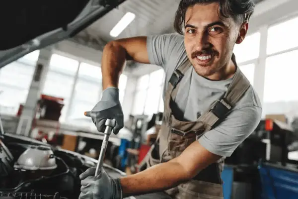 Mechanik w ogrodniczkach i rękawiczkach naprawia silnik samochodu kluczem nasadowym.