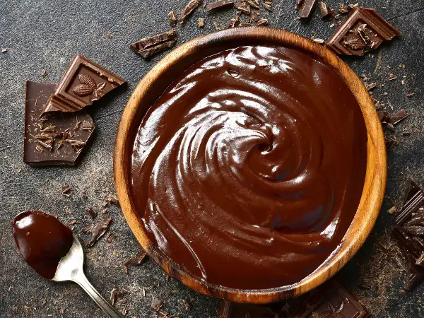 Un bol en bois déborde de chocolat fondu, prêt à travailler. Des éclats de chocolat et une cuillère en argent complètent cette scène gourmande.