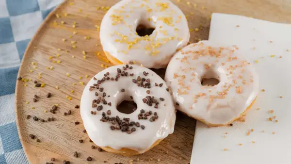 Drei Donuts mit weißer Glasur und Streuseln aus Schokolade, Zucker und Zitrone liegen auf einem Holzbrett.