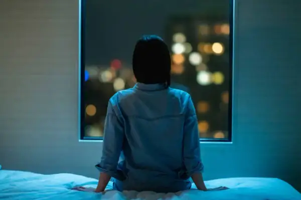 A person sits on a bed, looking out at city lights, feeling the weight of anxiety even when sleeping next to someone.