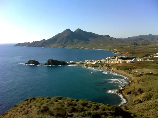 Vista panorámica de la cala del Toro, con sus aguas turquesas, rocas emergiendo y un pueblo blanco al fondo, bajo montañas verdes.