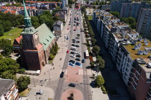 Widok z lotu ptaka na miasto z kościołem i długimi ogrodami. Ulicą jedzie autobus, a obok parkują samochody.