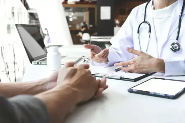 Lekarz omawia z pacjentem leczenie anty HCV. Na stole leżą tablet, laptop i butelka leków.