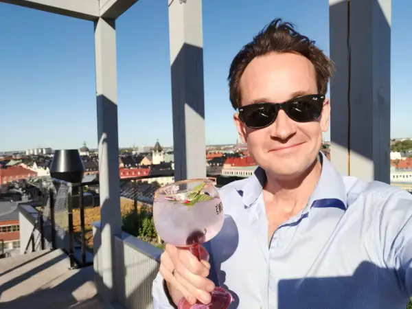 En man med solglasögon håller en rosa drink på en takterrass med utsikt över staden. En perfekt dag för en drink från arc blique by nobis.