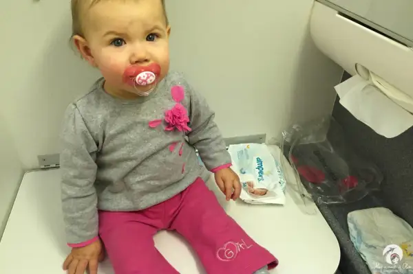 Mała dziewczynka z smoczkiem siedzi na siedzeniu w samolocie Ryanair. Obok niej leżą pieluchy i chusteczki.