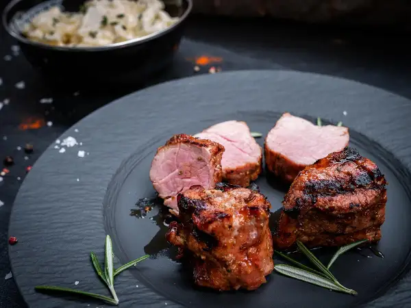 Saftige schweine medaillons, perfekt gegrillt, liegen auf einem dunklen Teller, garniert mit Rosmarin. Daneben eine Schüssel mit cremiger Beilage.