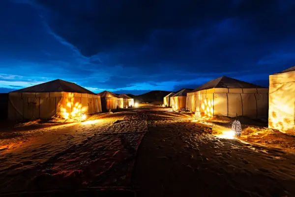 Campement de tentes illuminées dans le désert marocain, sous un ciel nocturne bleu profond. L'ambiance est magique.