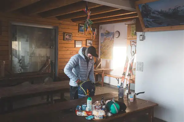 Dans une cabane de falaise, un skieur prépare son sac. Des skis, des cartes et des provisions sont visibles.