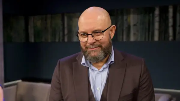 Szymon Bobrowski w garniturze i okularach, z brodą, wygląda jakby opowiadał o swoich filmach, serialach i programach.