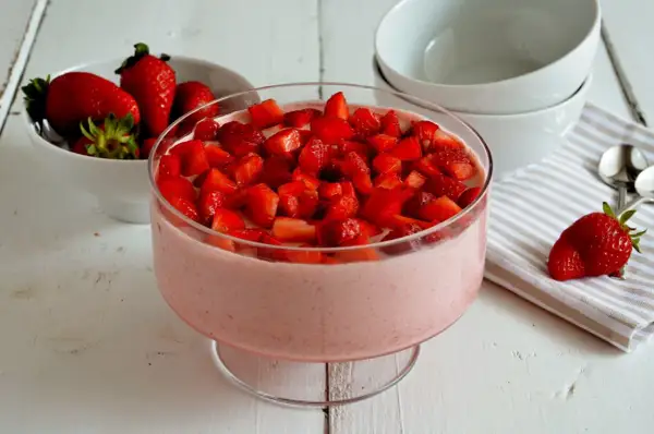 Delizioso mousse di fragole in una coppa di vetro, guarnito con fragole fresche a pezzetti. Accanto, una ciotola di fragole e ciotole bianche.