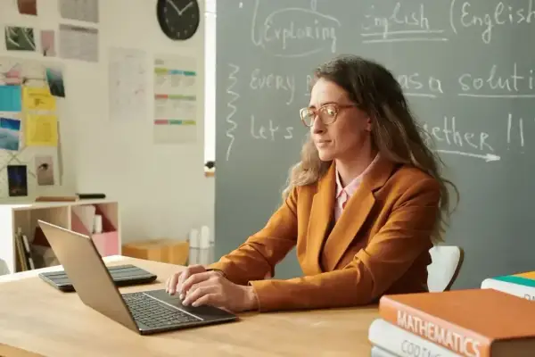Nauczycielka pracuje przy laptopie. Na tablicy widnieje napis "English". Karta oceny pracy nauczyciela może być wypełniana cyfrowo.