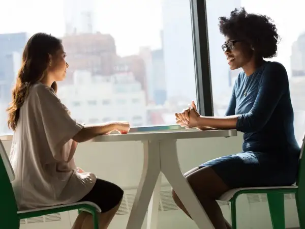 Dos mujeres conversan animadamente en una mesa junto a una ventana. Una de ellas, con gafas, sonríe mientras habla, demostrando su habilidad **speaking b1**.