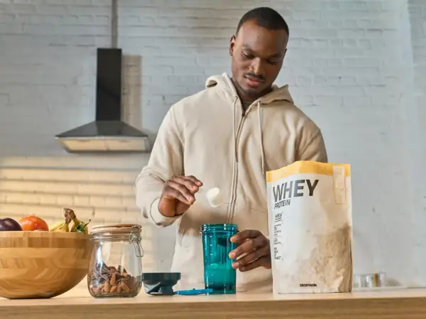 Mężczyzna przygotowuje odżywkę białkową po treningu. Na blacie stoi opakowanie WHEY PROTEIN, shaker i miska z owocami.