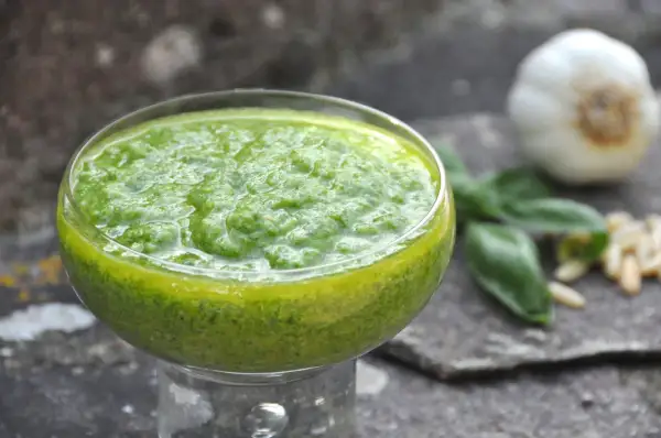 Pesto di basilico bimby in una coppetta, con aglio, pinoli e foglie di basilico sullo sfondo.