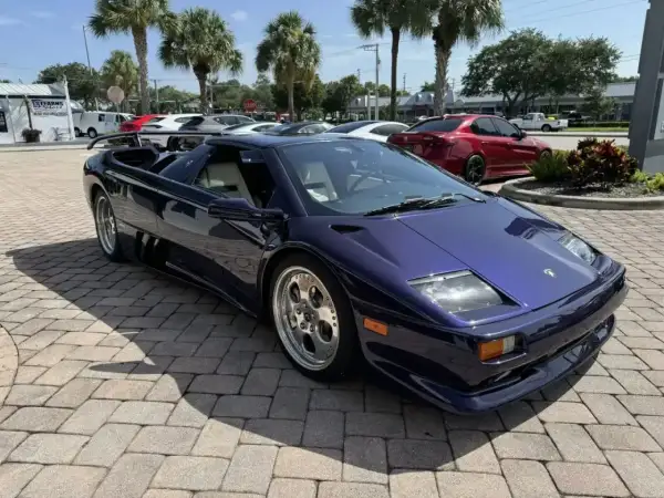 Fioletowy Lamborghini Diablo z jasnym wnętrzem, zaparkowany na brukowanym podjeździe, z palmami w tle.