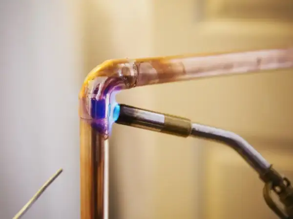 Un chalumeau chauffe un raccord coudé en cuivre pour le souder. La flamme bleue et violette illumine le métal.