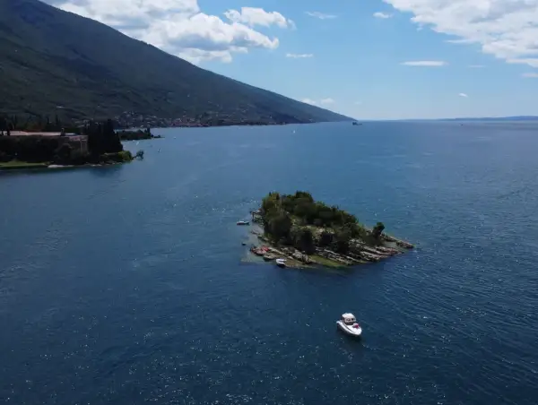 Un'isola verdeggiante nel Lago di Garda, con piccole imbarcazioni ormeggiate e una barca a motore che naviga nelle acque azzurre.