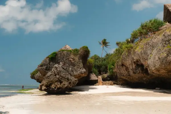 Zanzibar pora deszczowa. Skalista plaża z chatkami na klifie i palmą. W oddali widać samotną postać w czerwonej szacie.