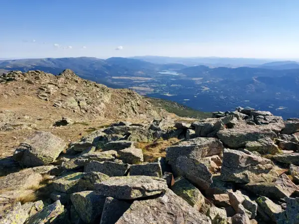 Vista panorámica de los picos de la Sierra de Guadarrama, con rocas cubiertas de líquenes en primer plano y un valle con un lago al fondo.