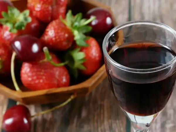 Szklanka ciemnego płynu, prawdopodobnie ratafia wódka, obok miski pełnej świeżych truskawek i wiśni.