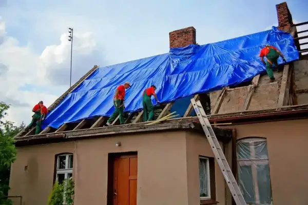 Pracownicy w zielonych ogrodniczkach i czerwonych koszulkach zabezpieczają dach niebieską plandeką podczas przebudowy domu.