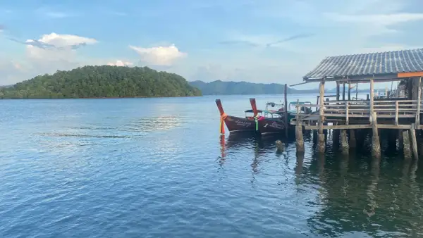Två longtailbåtar vid en träbrygga på Koh Jum. En grönskande ö syns i bakgrunden under en molnig himmel.