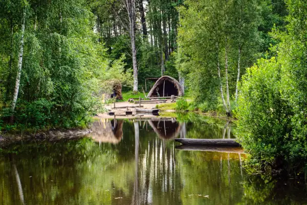 En stilla sjö speglar grönskande träd och en hydda vid Årås kvarn. En stockbåt vilar vid strandkanten.